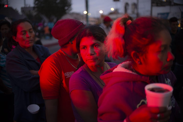 A woman looks out to the distance as a group of migrants, cast in a pink-hued light at night, wait for food.
